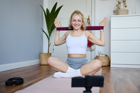 Portrait of young smiling woman, fitness instructor recording video about workout, showing how to exercise at home and use rubber resistance band, sitting on yoga matの写真素材