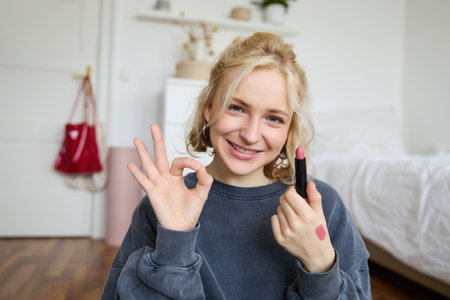 Portrait of young blond woman, content creator, recording video for social media about makeup, showing okay hand sign and lipstick, recommending good cosmetic productの写真素材