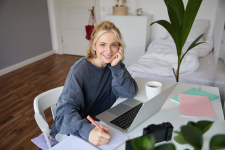Portrait of young woman distance learning, working from home with laptop, making notes, student studying on remote, doing online courseの写真素材