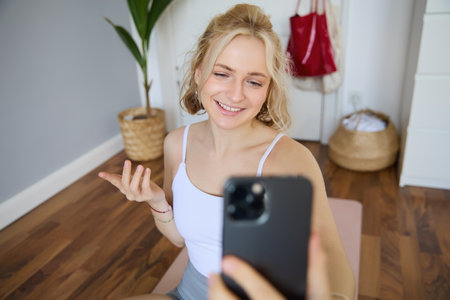 Portrait of beautiful young fitness trainer, showing home workout, taking selfie on mobile phone while sitting on yoga rubber mat in bedroom, wearing activewearの写真素材