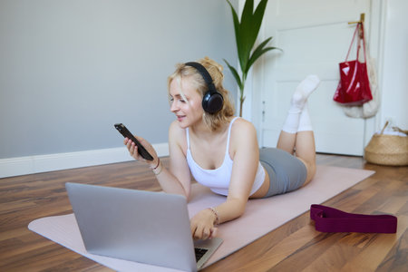 Portrait of young fitness woman, choosing workout on laptop, wearing headphones and using mobile phone exercise appの写真素材