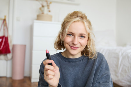 Portrait of smiling beautiful woman in her room, sitting and showing lipstick, recommending favourite beauty product, content maker recording a video of herself for social media blogの写真素材