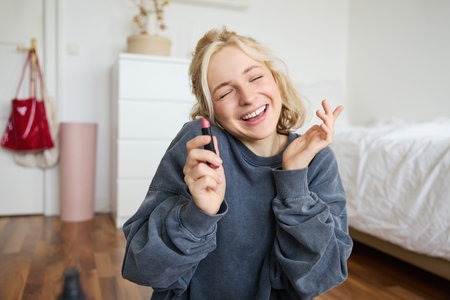 Portrait of smiling beautiful woman in her room, sitting and showing lipstick, recommending favourite beauty product, content maker recording a video of herself for social media blogの写真素材