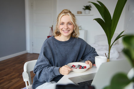 Portrait of young woman eating healthy meal in a room, watching videos on laptop, having lunch in front of computer, smiling and looking happy at cameraの写真素材