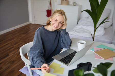 Portrait of smiling, beautiful blond woman, writing down notes, doing homework, studying from home, doing distance learning, online course, working remotely in her roomの写真素材