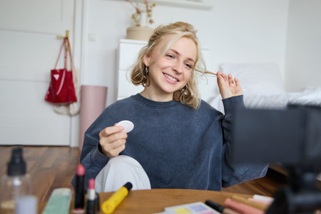 Portrait of smiling young woman applies makeup, shows beauty tutorial on video, records vlog on digital camera in her room, recommends cosmetic productsの写真素材