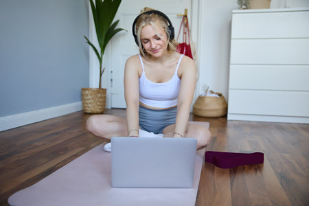 Portrait of smiling sporty woman at home, doing workout on rubber yoga mat, using laptop and wireless headphonesの写真素材