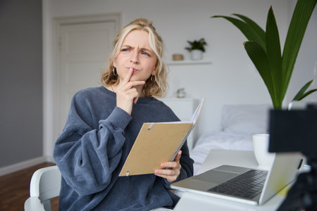 Portrait of young woman, student sitting in room, studying from home, remote education concept, holding notebook and thinking, looking aside with thoughtful face, concentrating on assignmentの写真素材