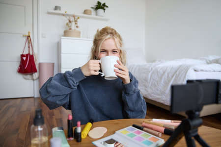 Image of young woman, makeup vlogger, sitting in bedroom with digital camera, drinking tea and talking, creating lifestyle video, social media contentの写真素材