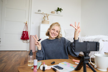Portrait of young blond woman, teenage girl records video for her social media account, shows makeup on camera, recommends lipstick to online followers, creates content in her roomの写真素材