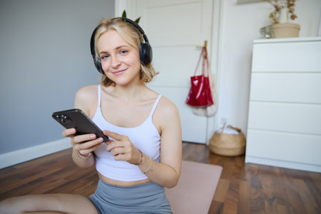 Close up portrait of athletic young woman, holding mobile phone and wearing wireless headphones, workout at home, using sport app during fitness trainingの写真素材