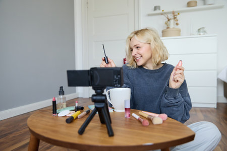 Portrait of young smiling woman in her room, recording video on camera, lifestyle vlog for social media, holding mascara, reviewing her makeup beauty products, showing how to use cosmeticsの写真素材