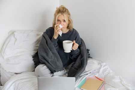 Portrait of young woman sitting at home sick, feeling unwell, drinking tea, spending time in bed, has runny nose, working on laptopの写真素材