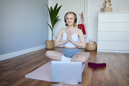 Portrait of blond young woman meditating, practise yoga, using yoga podcast to workout at home, sits on rubber matの写真素材