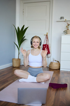 Vertical shot of smiling woman in wireless headphones, fitness instructor connects to online workout training session, shows exercises using laptop camera, sits on rubber yoga mat at homeの写真素材