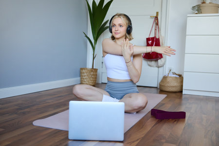 Portrait of beautiful, athletic woman at home, doing workout, watching fitness instructor video on laptop, wearing headphones, stretching her armsの写真素材