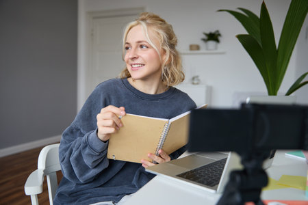 Portrait of smiling blond woman, sitting in bedroom, using laptop and digital camera, recording video for lifestyle blog, reading, using her notebookの写真素材