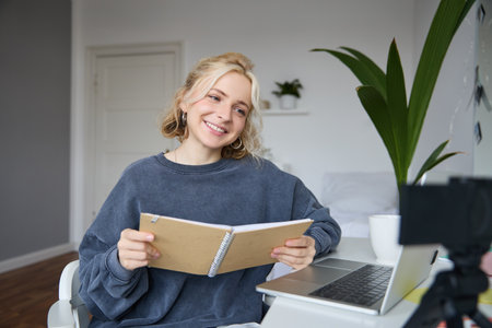 Portrait of smiling blond woman, sitting in bedroom, using laptop and digital camera, recording video for lifestyle blog, reading, using her notebookの写真素材