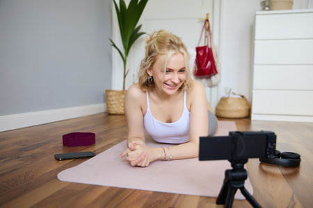 Portrait of young fitness instructor, vlogger creating content at home, doing workout and record exercises on digital camera, using rubber yoga matの写真素材