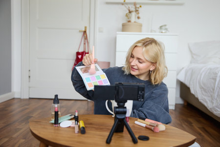 Portrait of smiling blond woman, teenage girl records a vlog on video camera, vlogger shows eyeshadows, tutorial how to put makeup on, sits in her room on floor near coffee tableの写真素材
