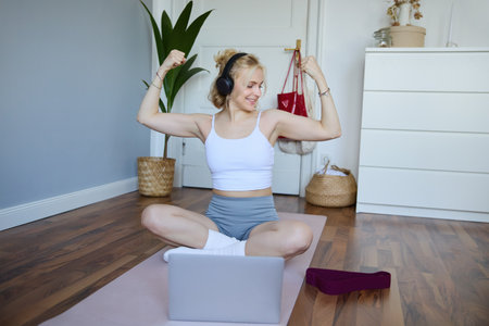 Portrait of young athletic woman doing workout at home, shows her muscles, strong biceps, sits on yoga mat and wears wireless headphonesの写真素材