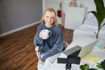 Portrait of young smiling woman, vlogger recording lifestyle video in her room, sits in front of laptop and digital camera, drinks tea, creates content for social media accountの写真素材