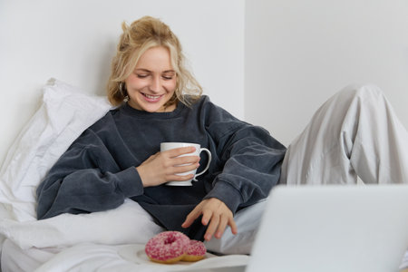 Portrait of happy blond girl, female model lying in bed in cosy clothes, eating and watching movie on laptop, smiling while looking at screenの写真素材
