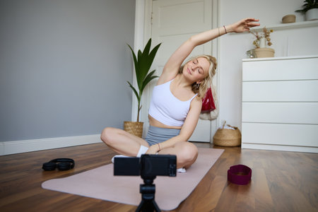 Portrait of young woman doing yoga at home, blogger recording video on digital camera while working out, personal fitness instructor showing exercises on rubber matの写真素材