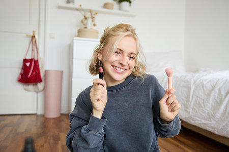 Woman beauty blogger, records video of herself sitting in a room and rating makeup products, puts on make up, holds lipstick and cosmetic brush in hand, using professional camera for content creationの写真素材