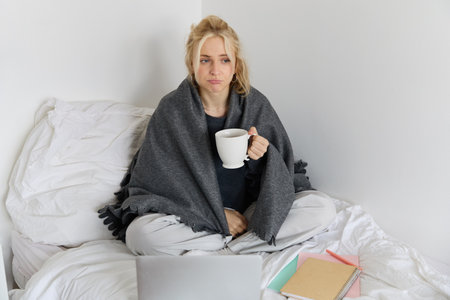 Portrait of woman catching a flu, sneezing, feeling sick, sitting on bed with laptop and working on remote while caught a coldの写真素材