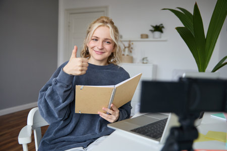 Smiling, positive young woman, shows thumbs up, sits in room with digital camera and laptop, records video, gives online tutorial, creates content for followersの写真素材