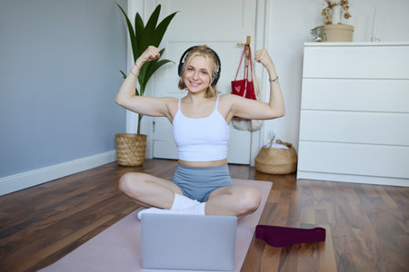Portrait of young happy woman, fitness blogger in wireless headphones, working out at home, sits on rubber mat with laptop and wireless headphones, shows her musclesの写真素材