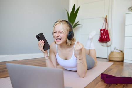 Portrait of young woman workout, watching exercise videos on laptop in headphones, lying on rubber mat with mobile phone and smilingの写真素材