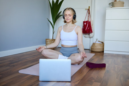 Portrait of young and relaxed woman using laptop and wireless headphones while meditating, practice yoga at home on rubber mat, sitting in asanaの写真素材