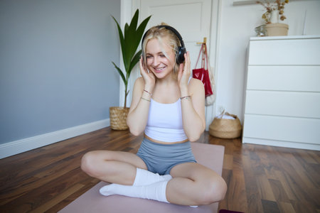 Sport and wellbeing concept. Smiling woman in wireless headphones, sitting on yoga mat, listens to music and workout at home, enjoys fitness training sessionの写真素材