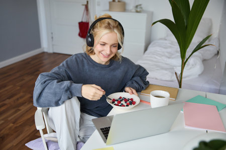 Portrait of happy young blond woman, sitting in a room, watching movie on laptop and eating healthy breakfast, drinking tea, resting on weekendの写真素材