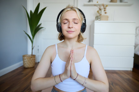 Portrait of woman meditating at home, sitting in headphones, listening to yoga podcast, holding hands in namaste signの写真素材