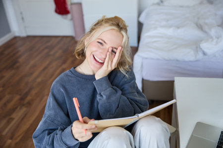 Portrait of happy, young blond woman, sitting in bedroom with notebook and pen, laughing and smiling, writing in journal, doing homework and smilingの写真素材