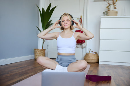 Portrait of young woman at home, connects to online gym training session, following fitness instructions on laptop, wearing wireless headphones during workoutの写真素材
