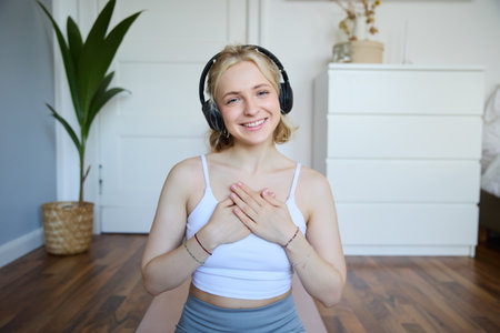 Portrait of woman feeling relaxed and in peace after meditation or yoga training at home, holding hands on chest, wearing headphones, smiling at cameraの写真素材