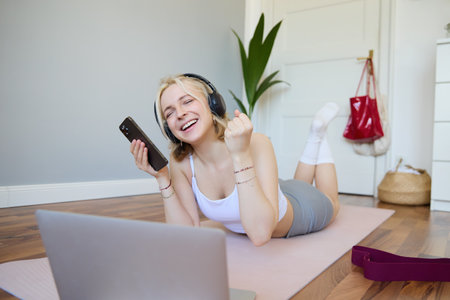 Young woman doing fitness workout at home, lying on rubber yoga mat, holding smartphone, wearing headphones, using laptop to watch sports videosの写真素材