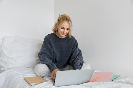 Image of happy young woman, student e-learning from home, connect to online course on her laptop, sits on bed with notebooks, smiling and looking happyの写真素材