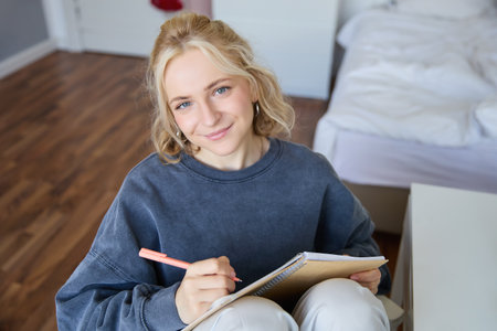 Close up portrait of beautiful young woman, writing in her diary, journal or planner, sitting in a room, smiling and looking at cameraの写真素材