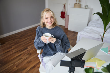 Image of young woman, social media influencer, editing her video on laptop, sits in a room with computer and digital camera, drinking coffee, smiling at cameraの写真素材