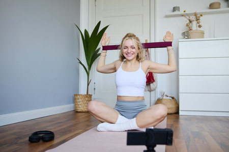 Portrait of young blond woman, social media vlogger using digital camera during workout, shooting video about exercises and fitness, stretching with rubber resistance bandの写真素材
