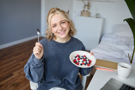 Portrait of smiling girl vlogger, blogger holding bowl with dessert, showing it on camera to audience, recording vlog in roomの写真素材
