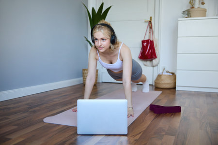 Portrait of young athletic woman in wireless headphones, standing in plank, following online workout...
