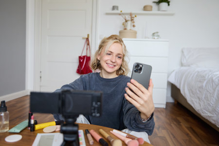 Portrait of young beautiful woman, lifestyle vlogger, sitting on bedroom floor, recording video on digital camera, using smartphone for online live stream, chatting with audience, reading messagesの写真素材