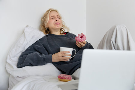 Portrait of sad and depressed woman, feeling heartbroken, lying in bed with comfort food, eating doughnut, crying from upsetting movie, watching something on laptopの写真素材