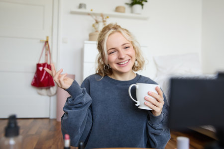 Portrait of cute smiling woman, blond girl drinks tea and records a casual, lifestyle video blog, vlogger sits in a room with camera and stabiliser, holds cup of coffee and talksの写真素材